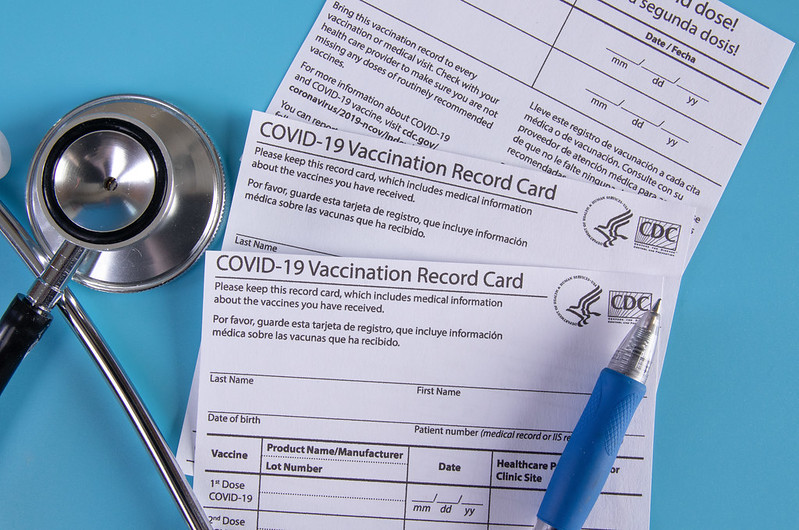 Three COVID-19 vaccination record card, stethoscope and pen on blue background.