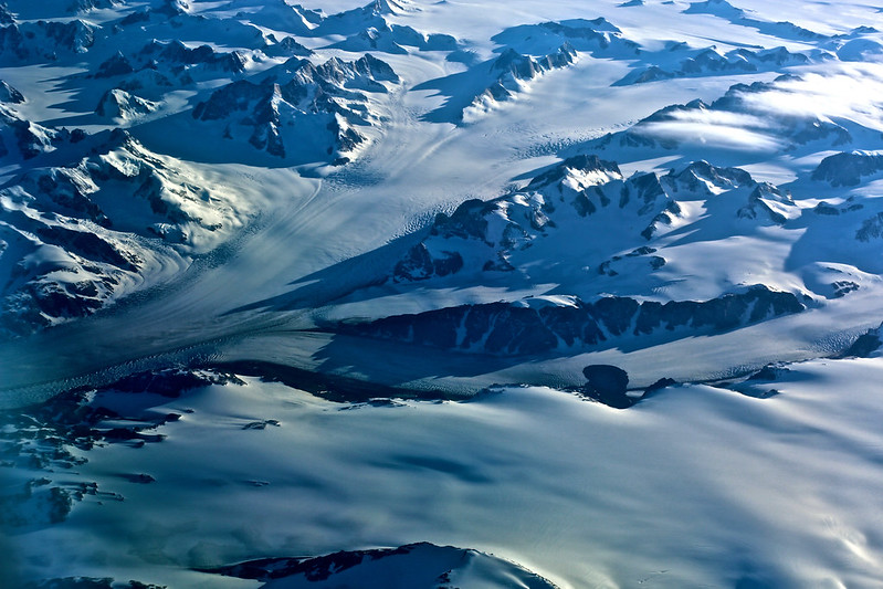 A portion of Greenlands frozen interior with a view of a glacier, the ice sheet and hill peaks seen from above.