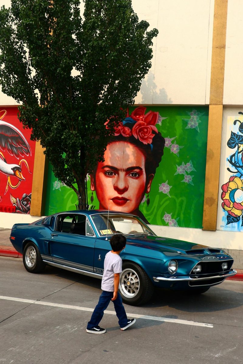 Young person walking by observing a classic car and a mural behind it of Frida Kahlo.