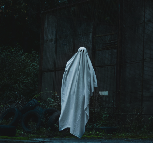 Someone in the dark in a ghost costume, with tires, bushes, and a fence in the background